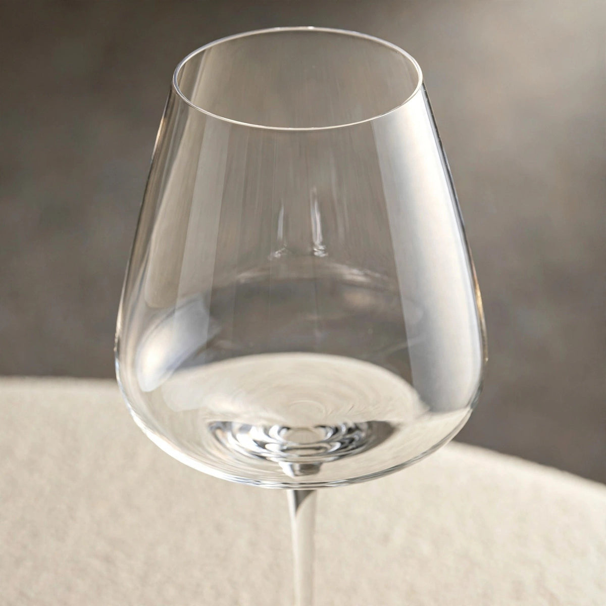A close-up shows an empty clear wine glass bowl and thin rim against a blurred gray background above a cream surface.