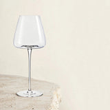 An empty clear wine glass with a long stem stands on a pale textured stone pedestal against a cream background.