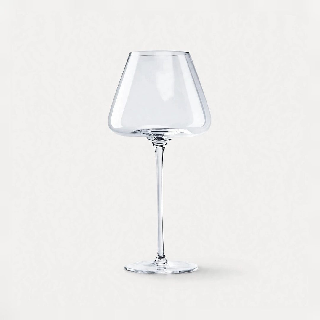 A single empty clear wine glass with an angular bowl and long stem stands centered against a pale background.