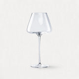 A single empty clear wine glass with an angular bowl and long stem stands centered against a pale background.
