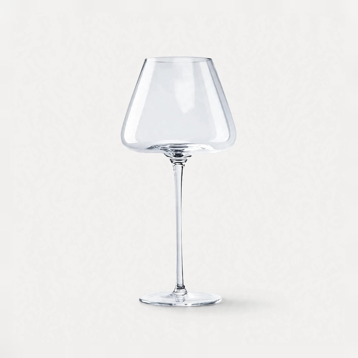 A single empty clear wine glass with an angular bowl and long stem stands centered against a pale background.