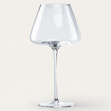 An empty clear wine glass with a broad angular bowl and tall slender stem stands against an off-white background.