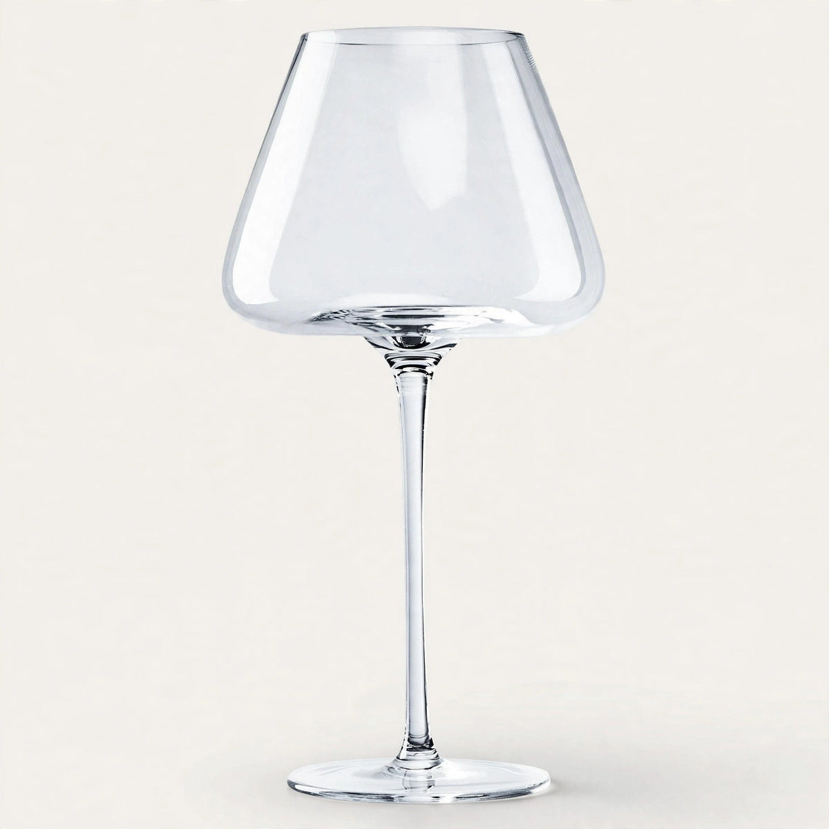 An empty clear wine glass with a broad angular bowl and tall slender stem stands against an off-white background.