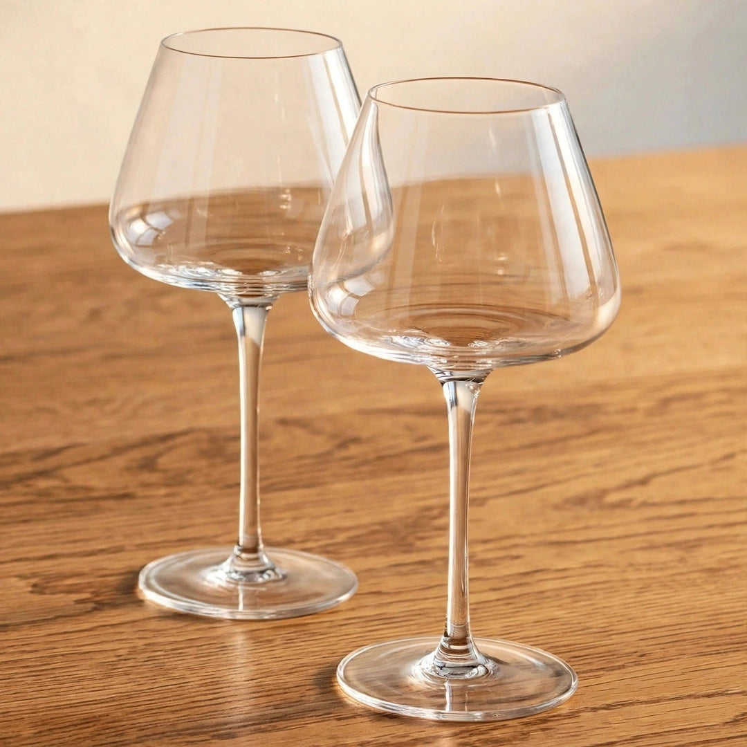 Two empty clear wine glasses with tapered bowls and tall stems stand diagonally apart on a wooden table.