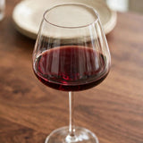 A clear wine glass partially filled with red wine stands on a wooden table with a blurred cream plate behind it.
