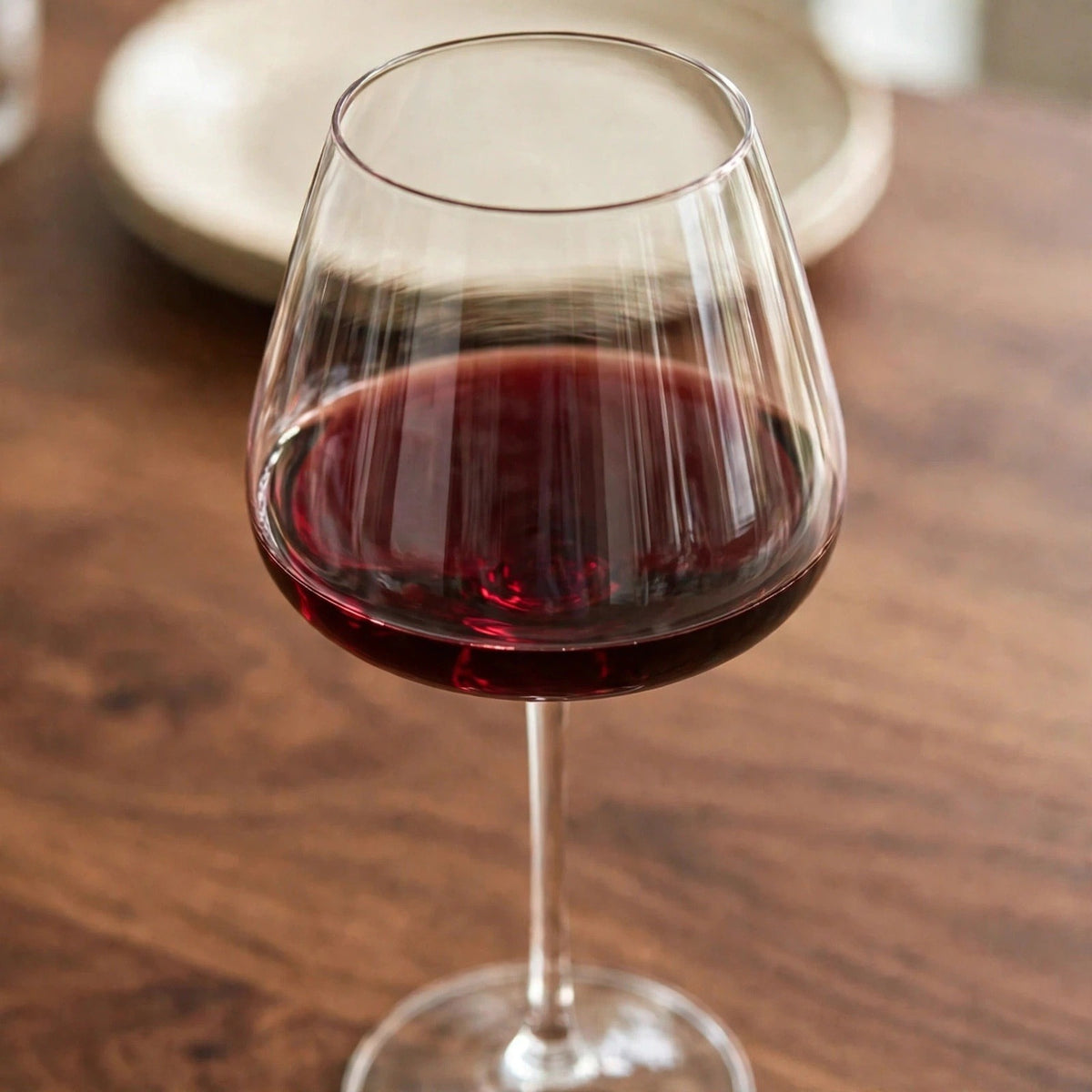 A clear wine glass partially filled with red wine stands on a wooden table with a blurred cream plate behind it.