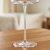 A close-up shows a clear wine glass's slender stem and circular base on a wooden table against a blurred beige interior.