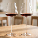 Two clear, long-stemmed wine glasses holding red wine stand on a sunlit wooden table with blurred chairs behind them.