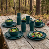 A set of dark teal green ceramic tableware, including bowls, plates, and cups, arranged on a rustic wooden picnic table outdoors with a blurred forest background.