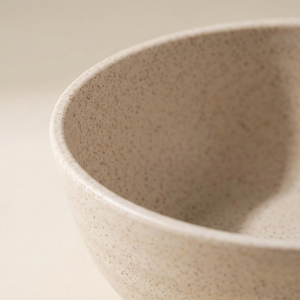 Close-up of a speckled ceramic bowl with a warm beige and cream glaze, showing the textured interior and rim against a matching neutral background.