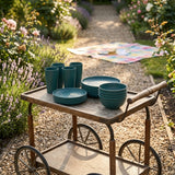 Dark green plates, bowls, and four tall cups sit on a wooden garden trolley beside flowers and a picnic blanket.