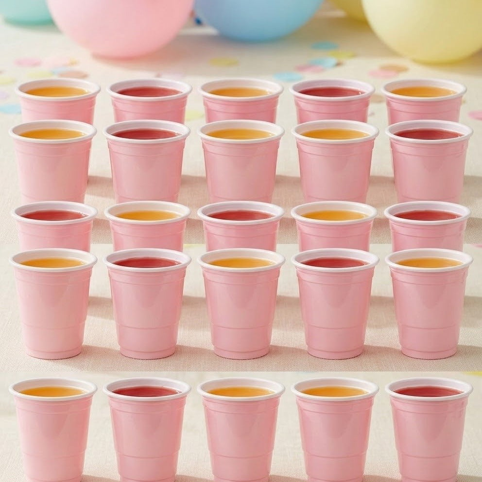 Pink cups with white rims hold red and orange drinks in rows on a pale table with pastel balloons behind.