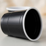 A black ridged cup with a white rim lies on its side on a pale surface, with another cup blurred behind.