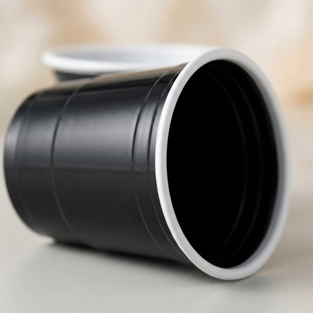 A black ridged cup with a white rim lies on its side on a pale surface, with another cup blurred behind.