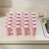 Twenty pink cups with white rims stand on a cream kitchen counter beside a sink, sponge, and drinking glass.