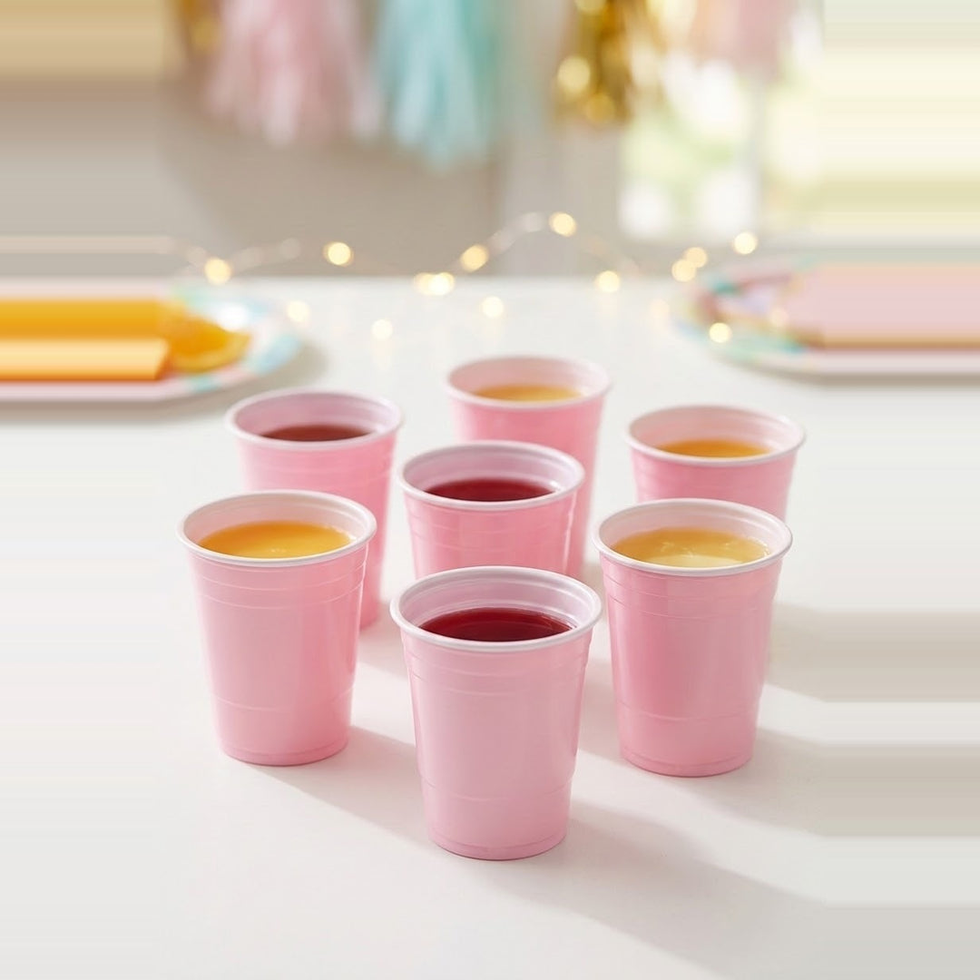 Seven pink cups with white rims hold red and orange drinks on a white party table with blurred decorations behind.