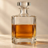 A clear square glass decanter with a broad stopper and heavy base stands in a warm studio setting.