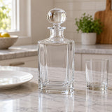 An empty square glass decanter stands beside a dinner plate and tumbler on a kitchen counter.