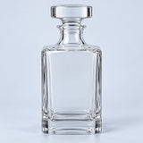 The clear smooth square glass decanter with its broad stopper stands against a pale blue backdrop.