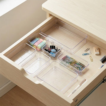 Clear plastic drawer organizer trays arranged inside an open light wood desk drawer, holding pens, clips, sticky notes, paper fasteners, and other stationery items.