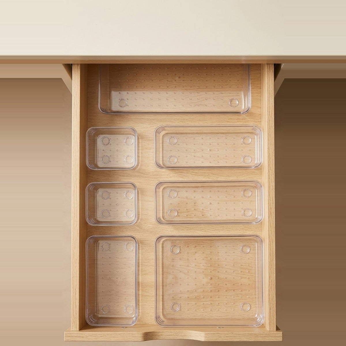 Six clear plastic drawer organizer trays arranged inside an open light wood drawer, shown against a warm beige background.
