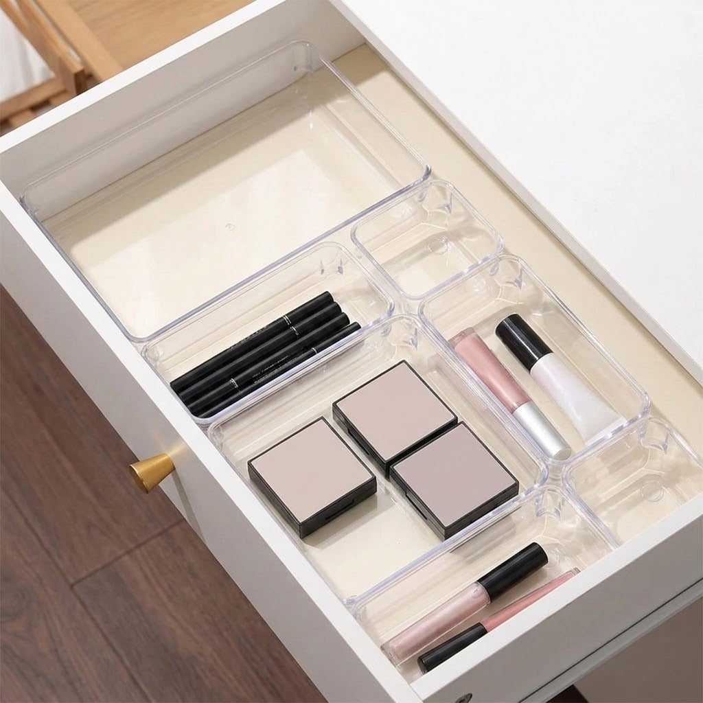 Clear plastic drawer organizer trays arranged inside an open white drawer, holding makeup items such as lipsticks, compacts, and a pencil. A wooden floor is visible at the bottom left.