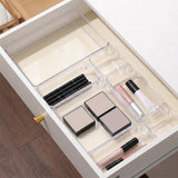 Clear plastic drawer organizer trays arranged inside an open white drawer, holding makeup items such as lipsticks, compacts, and a pencil. A wooden floor is visible at the bottom left.