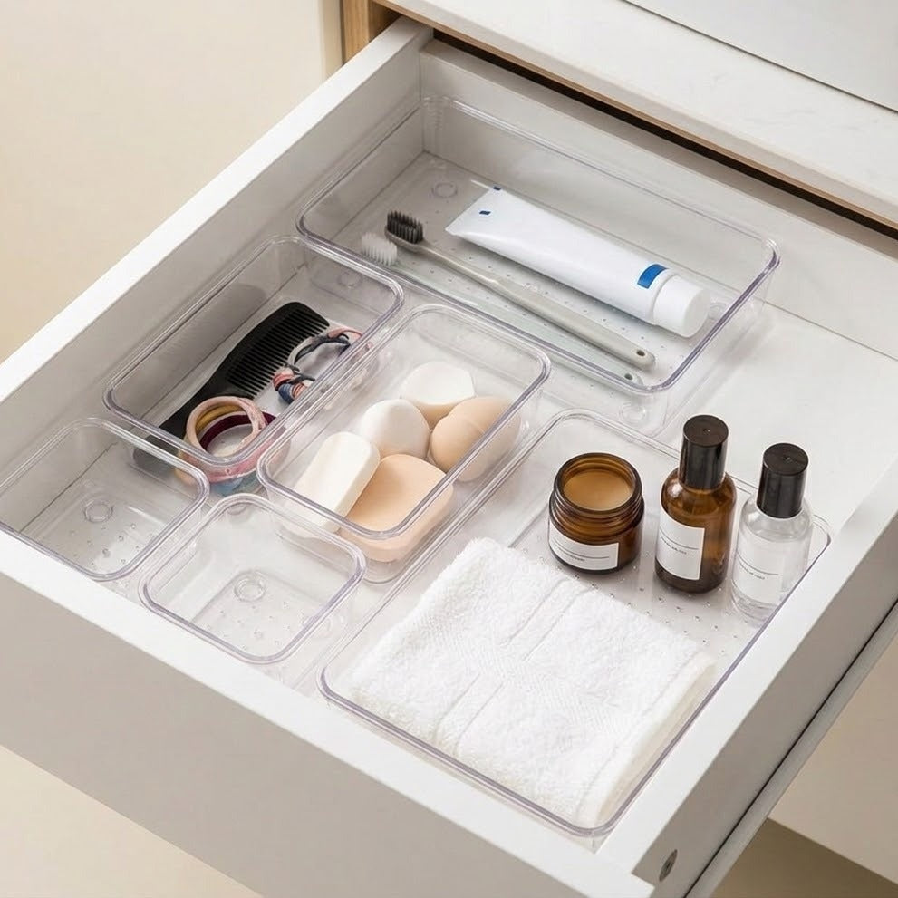 Clear plastic drawer organizer trays arranged inside an open white drawer, holding a toothbrush, tube, compact, makeup sponges, bottles, and a folded towel.