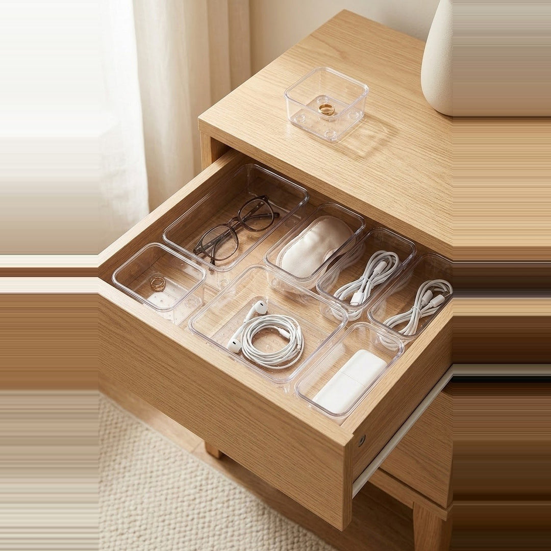 Clear plastic drawer organizer trays arranged inside an open wooden drawer, holding eyeglasses, a mouse, earphones, cables, and a small rectangular item. A matching clear tray sits on the wooden bedside table above.