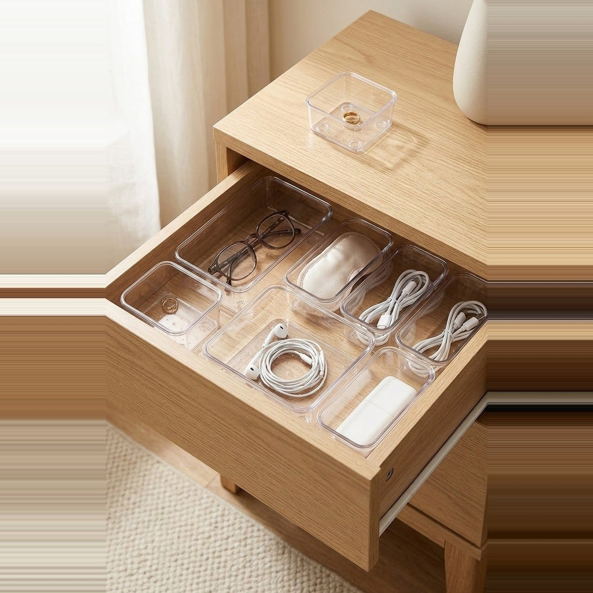 Clear plastic drawer organizer trays arranged inside an open wooden drawer, holding eyeglasses, a mouse, earphones, cables, and a small rectangular item. A matching clear tray sits on the wooden bedside table above.