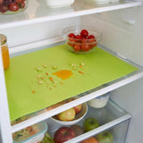 A bright green textured refrigerator shelf mat on a shelf inside an open refrigerator, surrounded by food items like tomatoes, apples, and juice.
