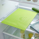 A bright lime green textured refrigerator mat placed on a glass shelf inside a refrigerator, with water droplets on top to demonstrate water resistance. A cucumber and a folded cloth are visible nearby.