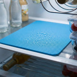 A blue textured silicone cutting mat with water droplets on top, shown on a refrigerator shelf with bottles and food containers in the background.