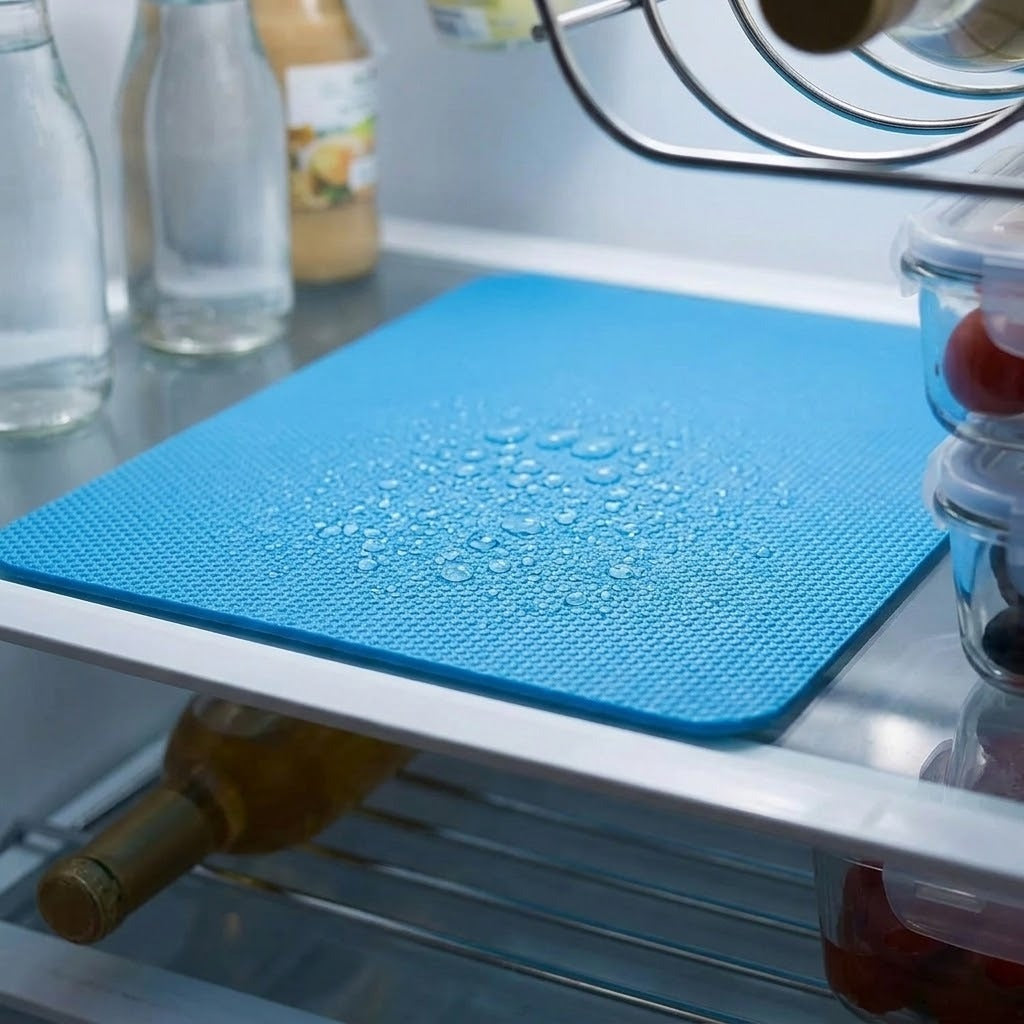 A blue textured silicone cutting mat with water droplets on top, shown on a refrigerator shelf with bottles and food containers in the background.