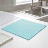 A square light blue silicone cutting board with a raised textured pattern sits on a white kitchen countertop near a sink. A clear plastic drying rack and folded beige towels are also visible in the scene.