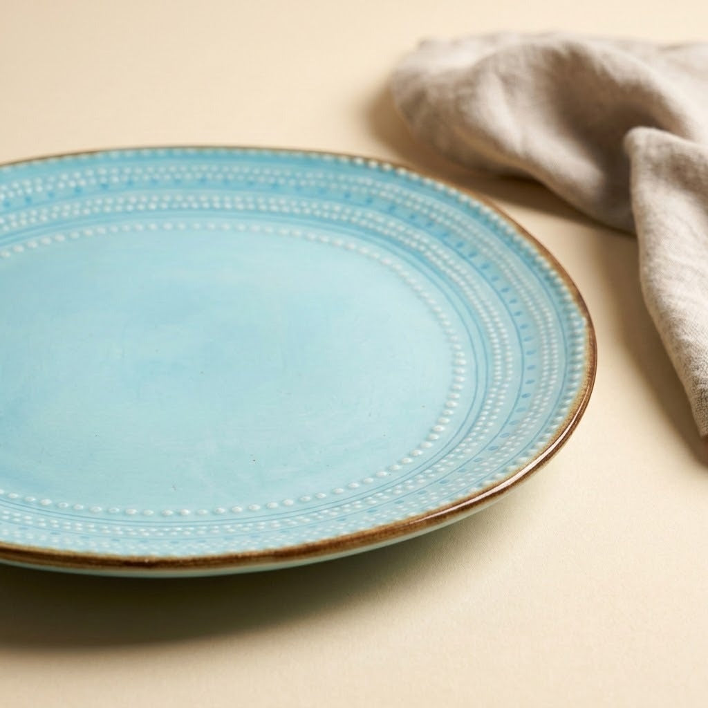 A light blue ceramic plate with a rustic brown rim and dotted border pattern, styled on a beige surface next to a beige linen napkin.