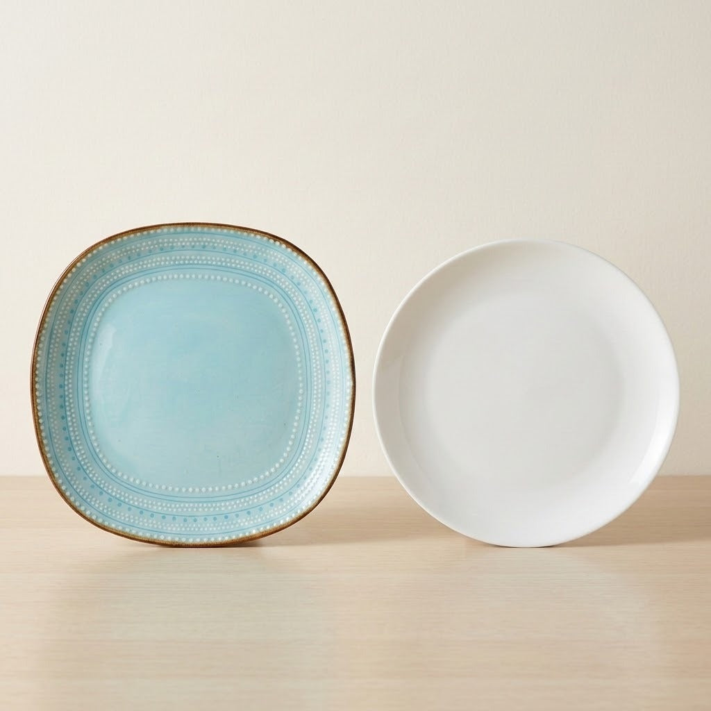Two ceramic plates on a light wood surface against a warm beige background: one square plate with a light blue interior, decorative dotted border, and brown rim, and one round white plate.