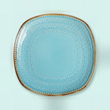 A square light blue ceramic plate with rounded corners, featuring concentric dotted patterns and a metallic gold rim, shown from a top-down view on a light mint green background.