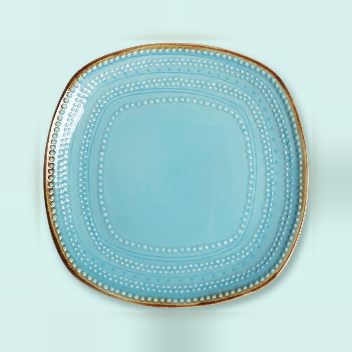 A square light blue ceramic plate with rounded corners, featuring concentric dotted patterns and a metallic gold rim, shown from a top-down view on a light mint green background.