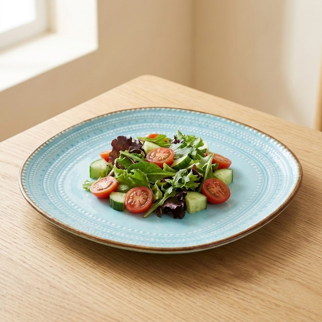 A light blue ceramic plate with a textured rim and gold-toned edge, holding a fresh green salad with tomatoes and cucumbers, set on a light wood table near a window.