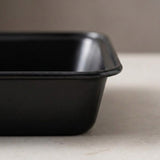A close-up of a matte black rectangular baking pan or tray with rounded corners, shown sitting on a light beige surface against a neutral gray background.