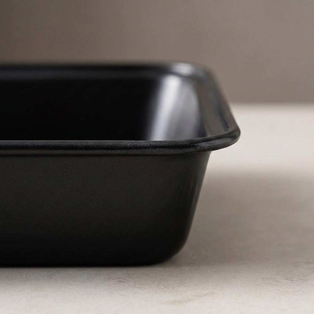 A close-up of a matte black rectangular baking pan or tray with rounded corners, shown sitting on a light beige surface against a neutral gray background.