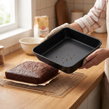 A person in a kitchen holds a black square baking pan over a cooling rack with a brownie on top. The scene includes a tiled wall, a window, bowls, and a bag of flour in the background.