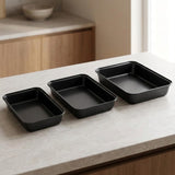 Three rectangular black baking pans of different sizes arranged diagonally on a light beige stone countertop in a modern kitchen setting.