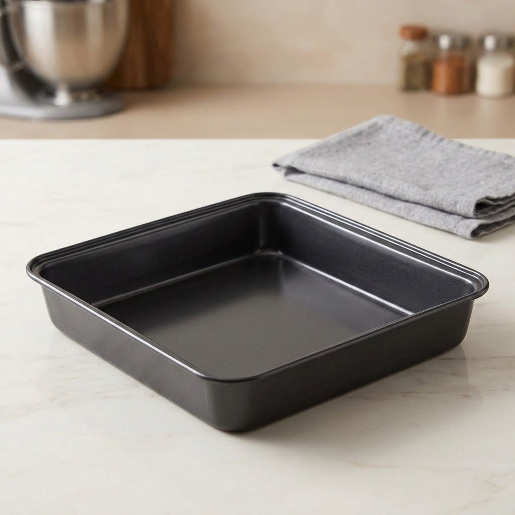 A square black non-stick baking pan with raised edges sits on a light countertop, with a folded gray cloth and blurred kitchen items in the background.