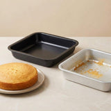 Two square baking pans on a light beige countertop: a black non-stick pan with a baked cake on a plate beside it, and a silver pan with crumbs inside.
