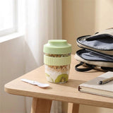 A reusable glass meal kit with a light green lid and textured green sleeve, filled with granola, yogurt, kiwi, and fruit pieces, sitting on a wooden table next to a spoon, notebook, and backpack.