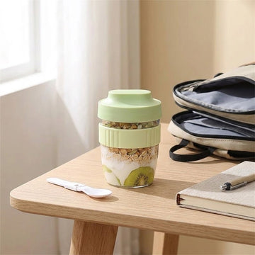 A reusable glass meal kit with a light green lid and textured green sleeve, filled with granola, yogurt, kiwi, and fruit pieces, sitting on a wooden table next to a spoon, notebook, and backpack.