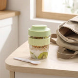 A light green portable meal prep container filled with granola, yogurt, and kiwi slices, sitting on a light wood table next to a white spoon and a beige tote bag in a bright kitchen setting.