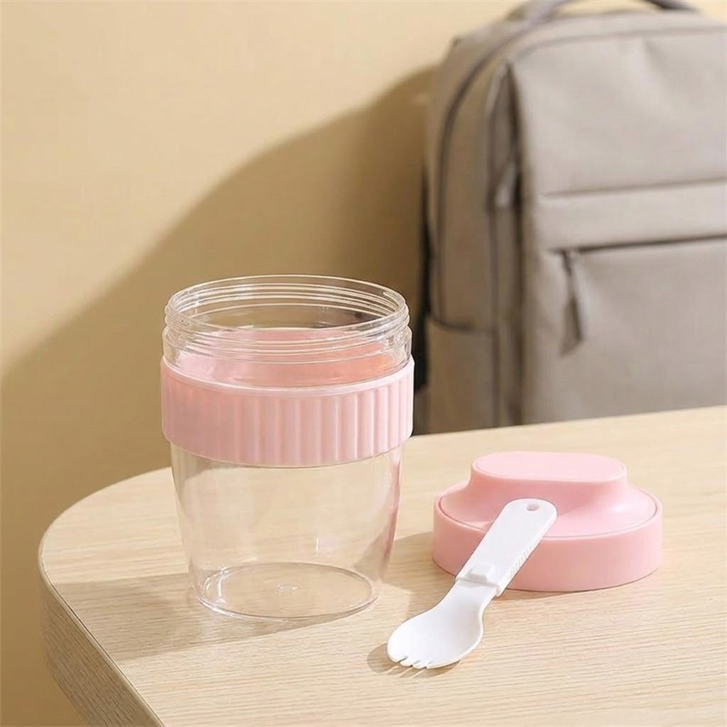 A clear plastic travel tumbler with a pink ribbed lid and matching pink cap, shown on a light wood table with a small white spoon. A gray backpack is blurred in the background against a beige wall.